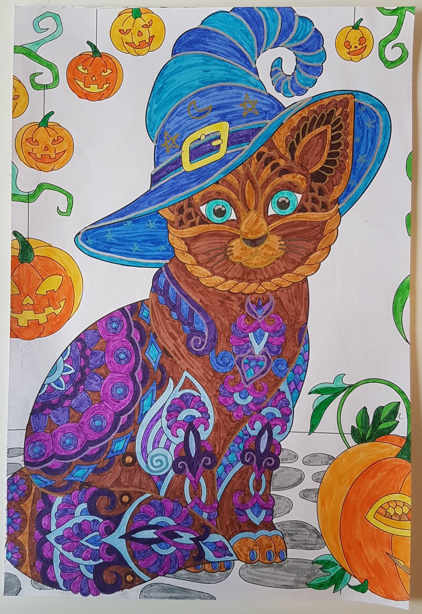 Cat with brown body fur, wearing a blue witch's hat, with artistic purple markings on its fur, and small orange punpkins in the background